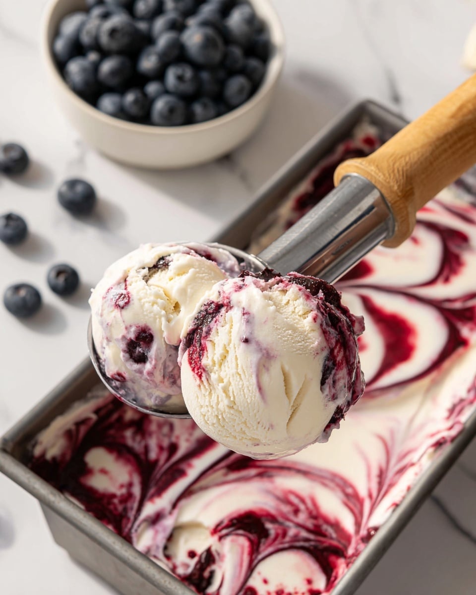 A close-up of a metal ice cream scoop holding two scoops of creamy white ice cream swirled with dark red berry sauce, showing a marbled pattern inside the scoop. Below, the same ice cream and berry sauce mix fills a rectangular metal container, where the swirling of white and deep red creates smooth, curved patterns. In the background, a white bowl full of fresh blueberries sits on a white marbled surface, and a woman's hand grips the wooden handle of the scoop. The scene is brightly lit with soft shadows, emphasizing the creamy texture of the ice cream and glossy berry swirls. photo taken with an iphone --ar 4:5 --v 7