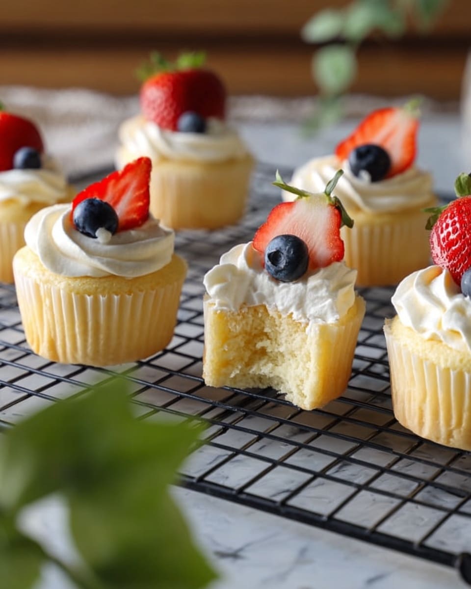 A group of small cupcakes sits on a black cooling rack placed on a white marbled surface. Each cupcake has one soft, light yellow cake layer topped with a generous swirl of fluffy white cream. On top of the cream, there is a fresh half strawberry along with a single round blueberry next to it, adding red and blue colors to the top layer. One cupcake in the foreground has a bite taken out of it, showing the soft, airy texture of the cake inside and the thick layer of cream beneath the fruit. A blurred green plant slightly covers part of the left side of the image. Photo taken with an iphone --ar 4:5 --v 7