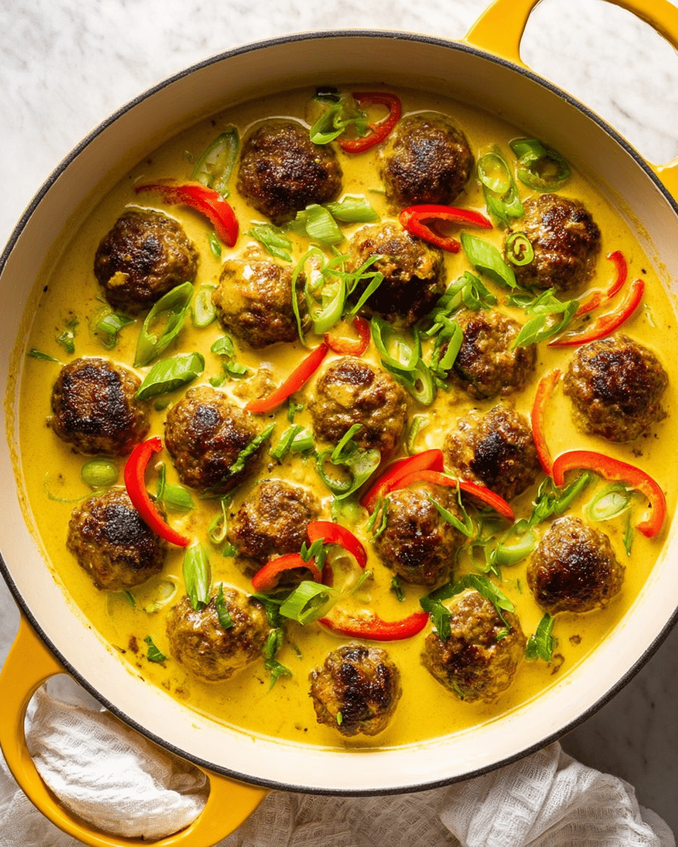 A white pan with yellow handles is filled with about twenty browned meatballs sitting in a thick, creamy yellow curry sauce. Thin slices of red bell pepper and bright green chopped scallions float on top, adding contrast and freshness. The surface beneath the pan has a white marbled texture, and a white cloth peeks out from underneath the pan's bottom edge. photo taken with an iphone --ar 4:5 --v 7