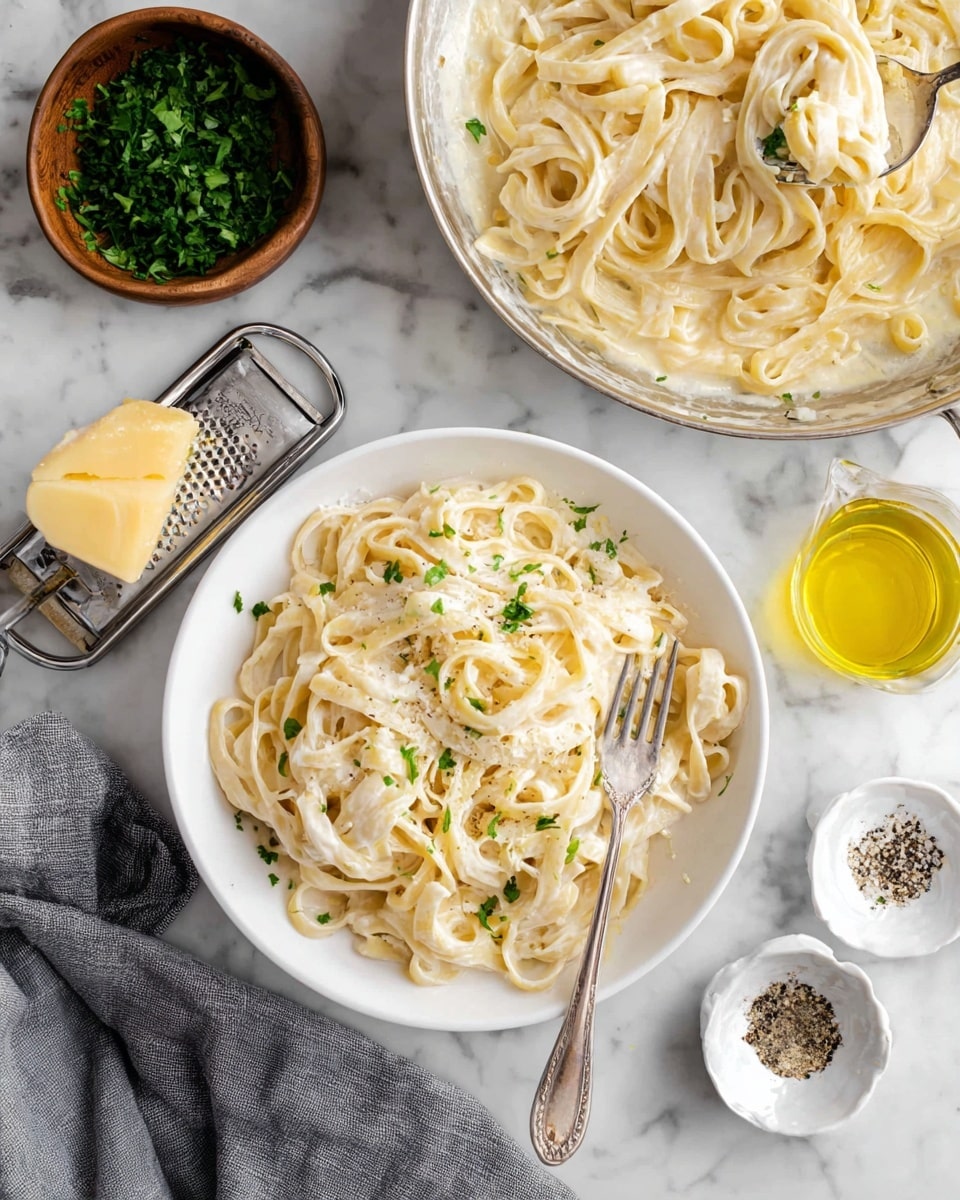 The image shows a white bowl filled with creamy fettuccine Alfredo pasta. The pasta is pale yellow with a smooth, thick white sauce coating each noodle. Small pieces of green parsley are sprinkled on top, adding spots of color. On the right side inside the bowl, a silver fork rests on the pasta, slightly lifting some noodles. Above the bowl, a silver pan holds more creamy fettuccine Alfredo with the same sauce and noodles twisted together. To the left, a small wooden bowl contains chopped green herbs, and next to it, a cheese grater with a yellow chunk of cheese rests on the white marbled surface. Also shown are two forks and two small white bowls with black pepper inside, along with a small glass container filled with yellow oil. A gray cloth is partially visible in the lower left corner. All of this is placed on a white marbled texture. photo taken with an iphone --ar 4:5 --v 7