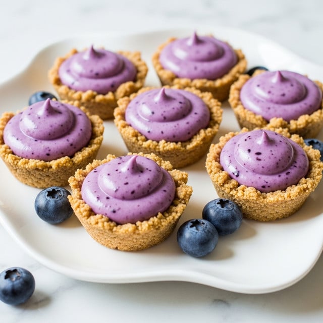 Seven small tartlets sit on a white plate with a soft, curved edge, placed on a white marbled surface. Each tartlet has a crumbly, light brown crust that looks textured and slightly rough. Inside each crust is a thick, creamy purple filling that is smooth with tiny darker specks, giving it a soft, whipped look. Around the tartlets, three fresh blueberries add a round, deep blue touch to the scene. The lighting brightens the colors and soft shadows keep the focus on the tartlets. photo taken with an iphone --ar 4:5 --v 7