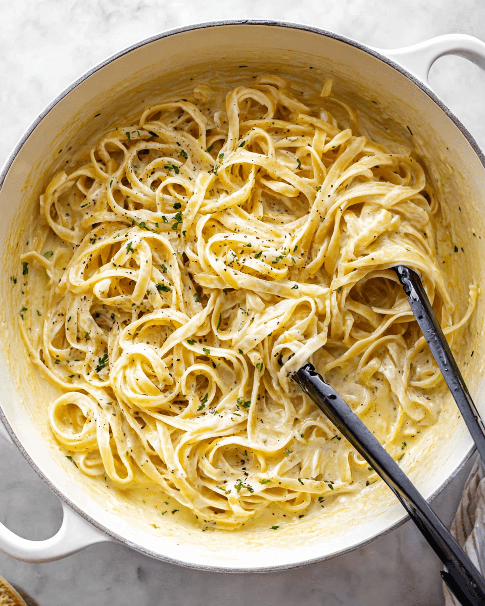 A white pot filled with creamy fettuccine pasta coated in a light yellow sauce, with visible specks of black pepper and small bits of green herb scattered throughout; the pasta is soft and slightly twisted in thick, smooth layers, some twirled around black and silver tongs resting on the right side of the pot. The pot sits on a white marbled textured surface, with a touch of shadow beneath the handle. photo taken with an iphone --ar 4:5 --v 7