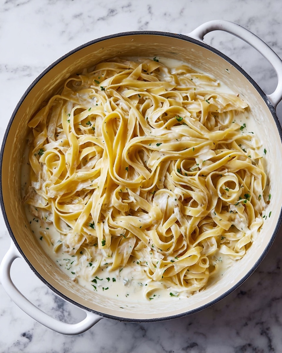 A white pot filled with creamy white sauce lightly speckled with green herbs forms the base layer, topped by a thick layer of long, flat, yellowish fettuccine noodles arranged loosely in a circular shape, some overlapping and twisting naturally. The creamy sauce has a smooth texture and small pieces of onion mixed in, visible around the edges under the noodles. The pot is on a white marbled surface. photo taken with an iphone --ar 4:5 --v 7
