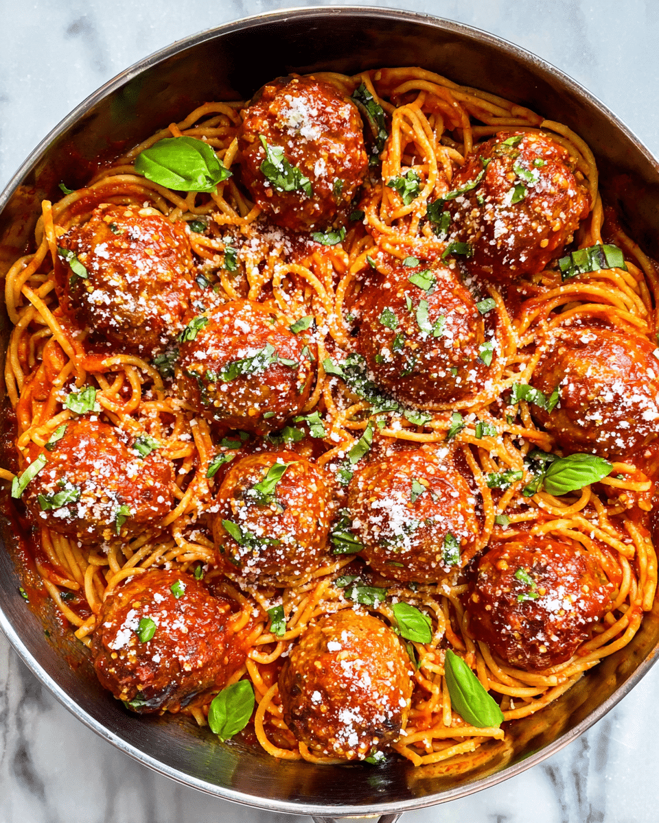A round pan filled with a base layer of orange-red spaghetti noodles covered in a rich tomato sauce, topped with nine large, glossy meatballs coated in the same sauce. The meatballs are sprinkled with small green herb pieces and white grated cheese. Fresh bright green basil leaves are scattered among the meatballs and noodles, adding a fresh touch to the dish. The pan is placed on a white marbled surface. photo taken with an iphone --ar 4:5 --v 7