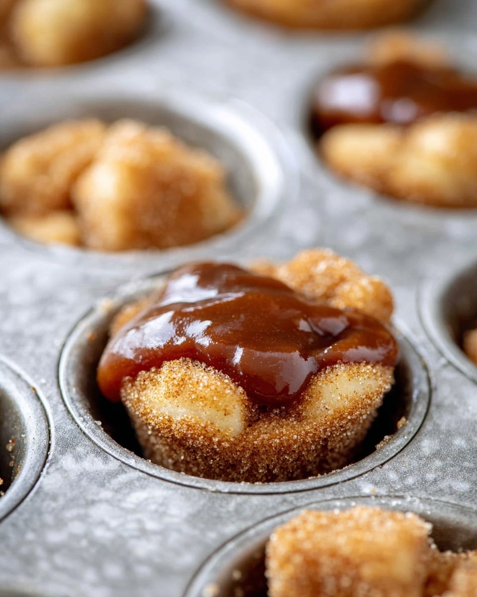 A close-up view of a single muffin cup in a silver metal muffin tray filled with bite-sized, light brown pieces coated in cinnamon sugar at the bottom, topped with a thick, glossy layer of dark brown sticky sauce covering the center. The muffin cup’s silver texture contrasts with the soft, grainy texture of the cinnamon sugar bites and the smooth sauce on top. The background shows parts of other similar muffin cups slightly out of focus, and all is set against a white marbled texture. photo taken with an iphone --ar 4:5 --v 7