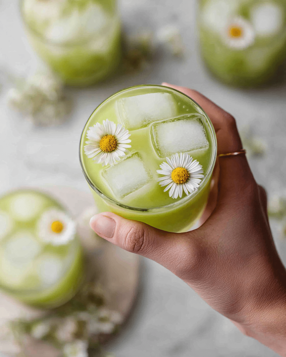 A close-up view of a glass held by a woman's hand filled with a light green drink, containing several clear ice cubes floating on top. Three small white daisies with yellow centers float gently on the surface of the drink, adding a delicate touch. In the blurred background, there are more glasses with the same drink and daisies on a white marbled surface, creating a soft and fresh atmosphere. photo taken with an iphone --ar 4:5 --v 7