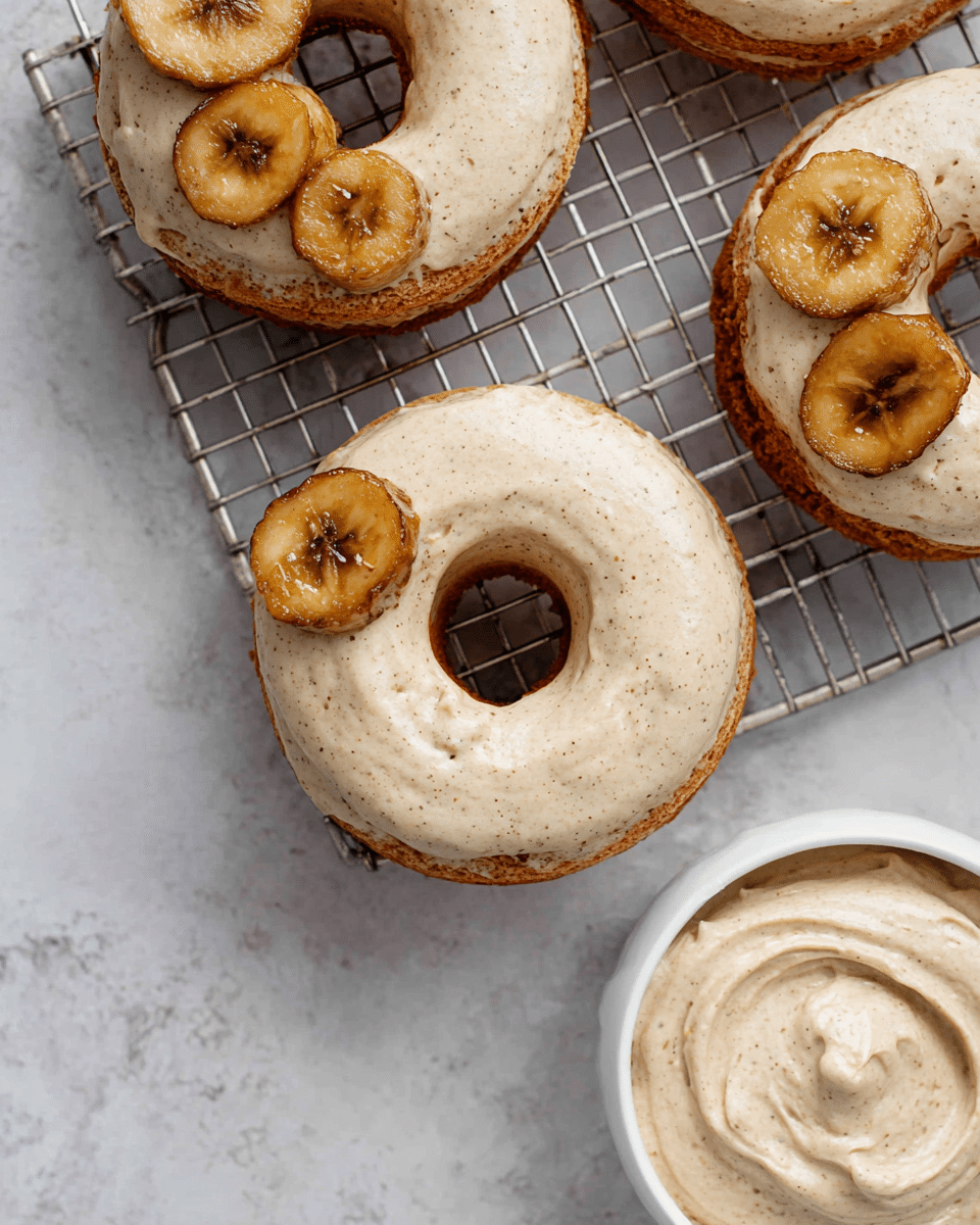 The image shows three donuts with a creamy beige frosting that has tiny brown specks, evenly covering the top surface of each donut. Each donut has three slices of golden-brown caramelized banana placed in a small group on one side near the hole in the center. Beneath the donuts, there is a silver cooling rack on top of a white marbled texture. In the lower right corner, there is a small white bowl filled with the same creamy frosting that is smooth with a slight swirl pattern. photo taken with an iphone --ar 4:5 --v 7