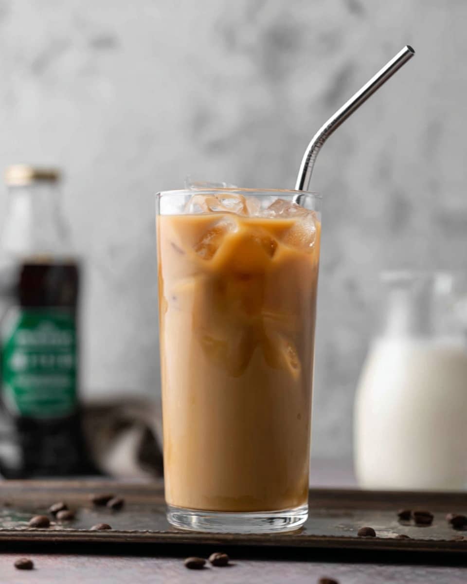 A tall clear glass filled with a light brown iced coffee drink showing a smooth, creamy texture with ice cubes floating just below the surface, a shiny silver metal straw bent at the top is placed inside the glass on the right side. The glass sits on a dark, slightly shiny metal tray scattered with a few coffee beans. The background is a soft, blurred white marbled texture with hints of a white creamer pitcher on the right and a bottle with a green cap, blurred on the left side. Photo taken with an iphone --ar 4:5 --v 7
