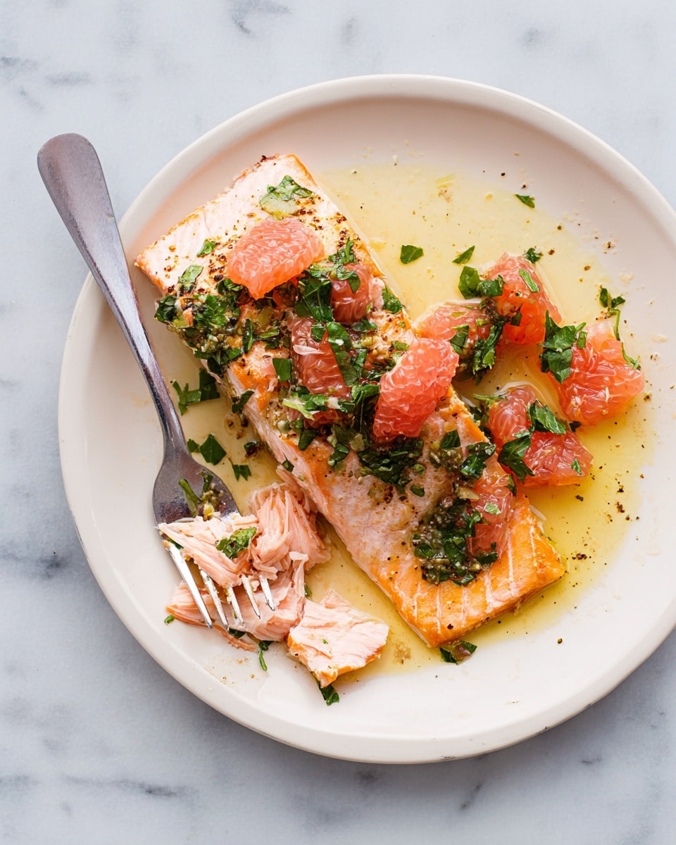 A white plate sits on a white marbled surface holding a cooked salmon fillet with a light pink-orange color and a slightly crispy, golden-brown edge on the right side. Over the salmon, there are chunks of deep pink grapefruit and bright green chopped herbs, scattered unevenly. A silver fork is placed on the left side of the plate, with some bits of salmon and herbs stuck to the tines. The dish appears fresh and lightly dressed with oil that adds a shiny texture. photo taken with an iphone --ar 4:5 --v 7
