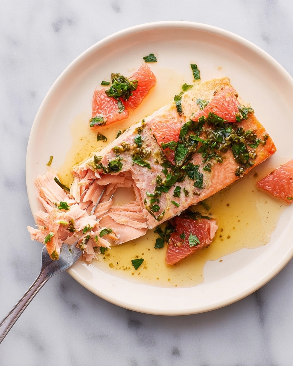 A white plate holds a piece of cooked salmon, its surface a light pink with a slightly browned edge and a moist, flaky texture visible where a piece has been cut. On top of the salmon and scattered around the plate are pieces of pink grapefruit and bright green chopped herbs, mixed with a shiny, light golden sauce. A fork with a piece of salmon on its tines rests on the left side of the plate. The plate is set on a white marbled surface. photo taken with an iphone --ar 4:5 --v 7