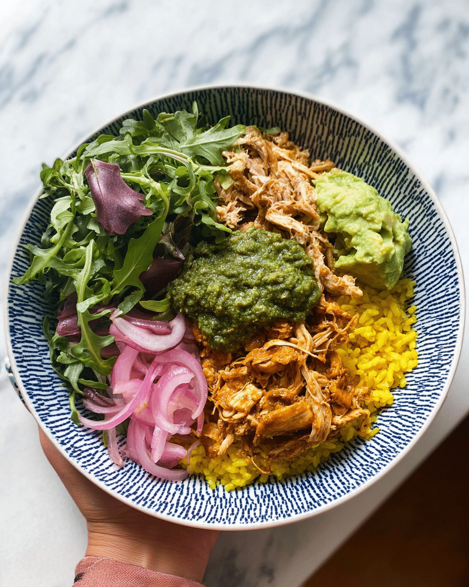 A white bowl with a blue geometric pattern holds a colorful layered dish. The bottom layer is bright yellow rice, followed by pulled shredded orange chicken on one side. Next to the chicken is a thick green pesto sauce with a coarse texture. Above this are thin slices of pink pickled onions, and a mixture of fresh green leafy veggies including arugula and purple-veined radish slices fills the remainder of the bowl. A scoop of mashed avocado sits near the greens, adding a creamy light green element. A woman's hand holds the bowl from the bottom left, showing painted nails. The background is a white marbled texture. photo taken with an iphone --ar 4:5 --v 7