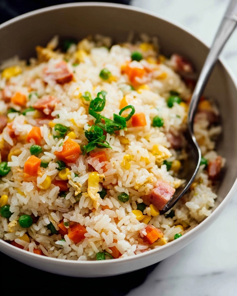 A close-up view of a bowl filled with fried rice, featuring fluffy white rice grains mixed with bright orange carrot cubes, small green peas, and yellow corn kernels. There are also small pinkish cubes of cooked ham scattered evenly throughout. Chopped green onions add a fresh green touch on top. A silver spoon is partially submerged in the rice, ready to scoop some up. The bowl is white, set against a white marble surface. photo taken with an iphone --ar 4:5 --v 7