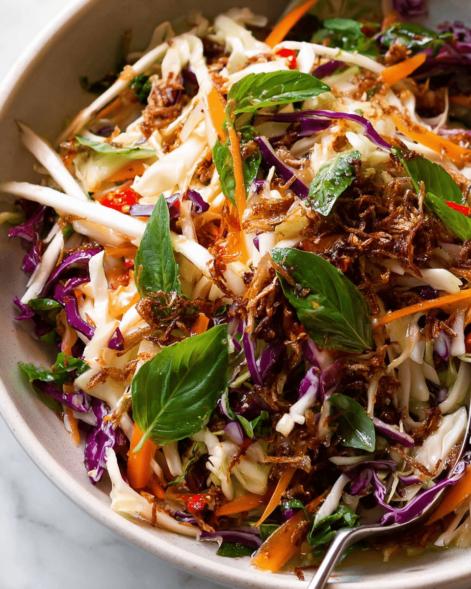 A close-up view of a fresh salad in a white bowl showing several colorful layers: thin strips of white and purple cabbage, thin orange carrot sticks, and bright green basil leaves scattered on top. The salad also has some crispy brown fried onions mixed within, adding texture. Some parts show a light dressing that makes the vegetables look shiny, with small red chili flakes visible. The bowl sits on a white marbled background, and a silver spoon rests inside the bowl on the right side. photo taken with an iphone --ar 4:5 --v 7