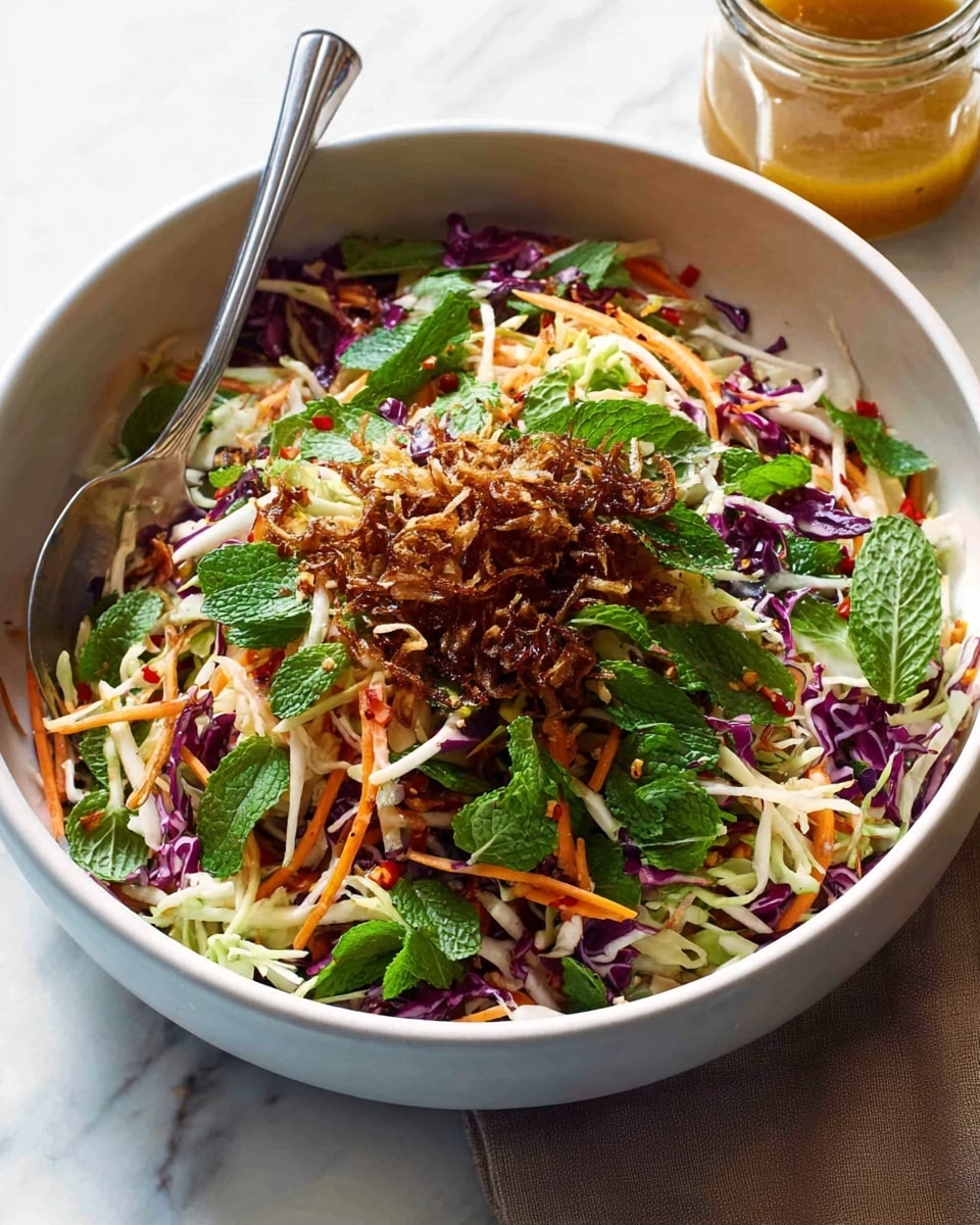 The image shows a white bowl filled with a colorful fresh salad. The bottom layer is a mix of thin, shredded white and purple cabbage, giving a crunchy texture. On top of this are thin, bright orange carrot strips scattered evenly. Fresh green herbs like mint and cilantro leaves are spread throughout, adding a bright green color. The salad is garnished with a small pile of crispy, brown fried shallots at the center. There are also some red chili flakes sprinkled lightly over the salad for a touch of red pop. A silver spoon rests inside the bowl on one side, and a glass jar with some kind of dressing is placed near the bowl on a white marbled surface. photo taken with an iphone --ar 4:5 --v 7