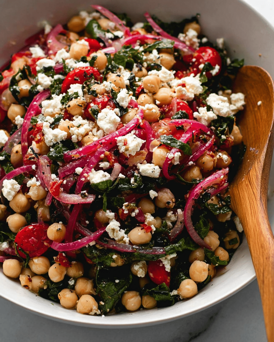 The image shows a close-up of a white bowl filled with a colorful salad made of multiple layers. The base consists of light beige chickpeas with a slight shine. Intermixed are chopped dark green leafy herbs and bright red grape tomato halves. Thin slices of reddish-pink pickled onions are scattered on top, adding light purple tones. Small crumbles of white feta cheese sit evenly across the salad, and some roasted red pepper strips add deeper red highlights. A wooden spoon is partially visible, stirring or scooping the salad from the right side, resting on a white marbled surface. photo taken with an iphone --ar 4:5 --v 7