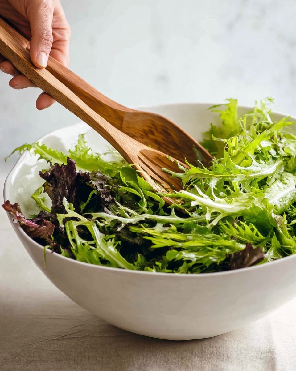 A white bowl filled with fresh mixed leafy greens, mostly light green with some darker green and purple leaves mixed in, showing a variety of textures with frilly edges and smooth leaves; two wooden salad servers, one held by a woman's hand and the other resting in the bowl, are tossing the greens gently. The background is a white marbled texture. photo taken with an iphone --ar 4:5 --v 7