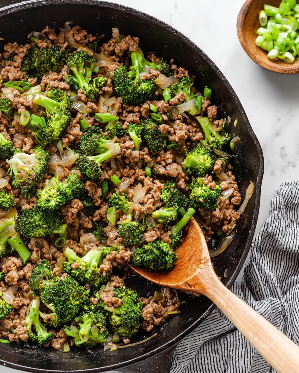 A close-up view of a black cast iron skillet filled with a stir-fry consisting of small, bright green broccoli florets, finely chopped translucent onions, and browned ground meat mixed in a light brown sauce. A wooden spoon rests inside the skillet, lifting a mix of broccoli and meat. The skillet sits on a white marbled surface, with a small wooden bowl of sliced green onions placed nearby, and a gray striped cloth partially visible in the corner. photo taken with an iphone --ar 4:5 --v 7