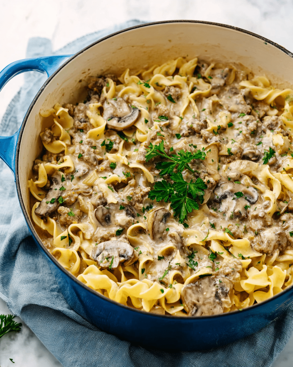 This image shows a close-up of a white cooking pot with a blue handle filled with creamy pasta. The dish has three layers: the bottom layer is wide egg noodles in a light yellow color, the middle layer includes sliced brown mushrooms, and the top layer is a creamy beige sauce mixed with small chunks of ground meat and sprinkled with finely chopped green parsley. There is a sprig of parsley placed on top for garnish. The pot is placed on a soft blue cloth on a white marbled surface. photo taken with an iphone --ar 4:5 --v 7