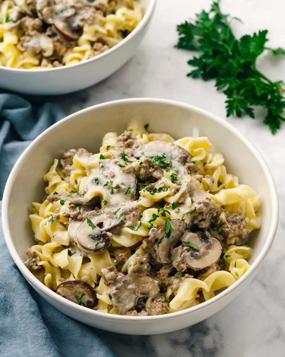 A white bowl filled with three layers of creamy pasta, each with a mix of light yellow noodles, browned ground meat, and sliced brown mushrooms coated in a thick, white sauce. The pasta looks soft and twisted with a slight sheen, while small green parsley pieces are scattered on top for color contrast. In the background, there is a second white bowl with the same pasta partly visible and a sprig of green parsley on the white marbled surface beside the bowl, with a soft blue cloth nearby. photo taken with an iphone --ar 4:5 --v 7