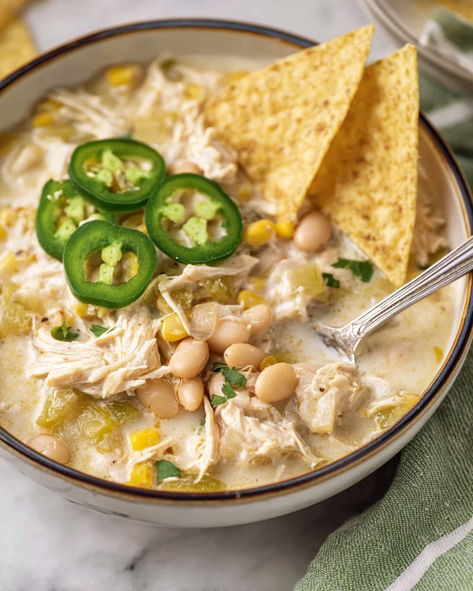A close-up of a bowl of creamy white chicken chili, with visible layers of shredded white chicken, pale yellow corn, light beige beans, and bits of green peppers all mixed in a thick white broth. Several bright green sliced jalapeños are placed on top on the left side. Two triangular light beige tortilla chips are stuck upright in the chili on the top right. A silver spoon is dipping into the chili on the right side, scooping some chili with chicken, beans, corn, and peppers. The bowl has a thin dark rim and is set on a white marbled surface with a green and white striped cloth partially visible on the right side. Photo taken with an iphone --ar 4:5 --v 7
