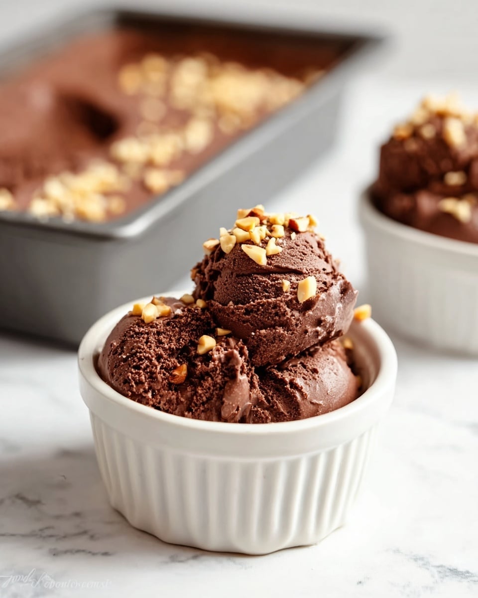A small white ceramic bowl holds three scoops of dark brown chocolate ice cream with a smooth yet slightly chunky texture, topped with light tan chopped nuts scattered unevenly on top. In the background, there is a close-up view of a rectangular baking pan filled with similar chocolate ice cream sprinkled with the same nuts, all set on a white marbled surface. Another bowl with chocolate ice cream and nuts is slightly visible to the right. The focus is sharp on the bowl in front, with a soft blur on the background items. Photo taken with an iphone --ar 4:5 --v 7