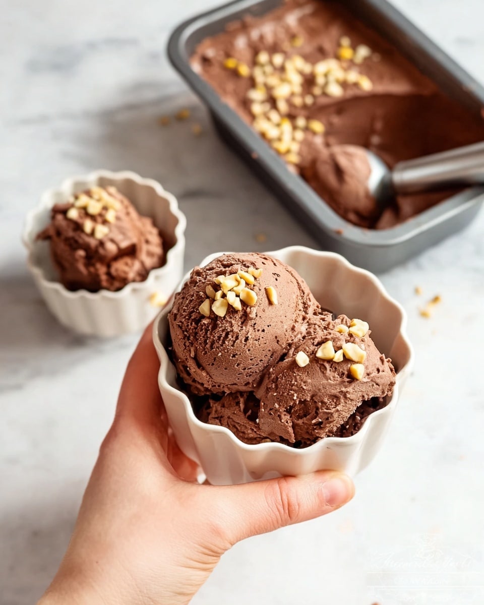 Two scoops of rich dark brown chocolate ice cream topped with small light brown nut pieces sit in a white ceramic bowl with a scalloped edge, held by a woman's hand in the foreground. Another bowl identical in shape and color, filled with the same chocolate ice cream and nut topping, rests on a white marbled surface next to it. In the background, a rectangular gray metal pan filled with more chocolate ice cream, some nut pieces scattered on top, and a scoop partially submerged, is also placed on the white marbled surface. The ice cream has a creamy and slightly textured look, with the nut pieces adding contrast on the smooth surface. Photo taken with an iphone --ar 4:5 --v 7