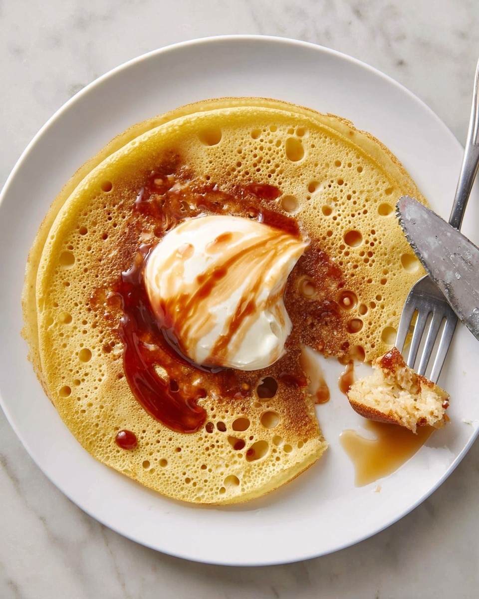 A single yellowish light pancake with many small holes covers the whole white plate, which sits on a white marbled surface; the pancake has a small cut piece lifted by a fork near the bottom right. On top of the pancake in the center, there is a dollop of smooth white cream, partially covered by a thick, shiny swirl of red-brown syrup. Some lighter syrup is drizzled over the cream and around the plate edges, and a metal knife with syrup drops rests on the right side of the plate. photo taken with an iphone --ar 4:5 --v 7