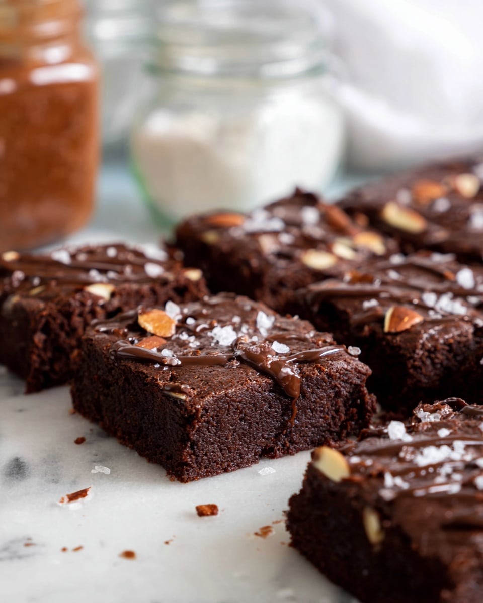 A close-up view of several square chocolate brownies placed on a white marbled surface, each brownie showing a rich, dark brown color with a slightly glossy top layer covered in chopped almond pieces and a drizzle of melted chocolate. The brownies are cut into thick squares, revealing a dense and moist texture inside. Small flakes of sea salt are scattered on top, adding a slight contrast to the dark surface. In the background, blurred glass jars with ingredients are visible, enhancing the homemade feel. photo taken with an iphone --ar 4:5 --v 7