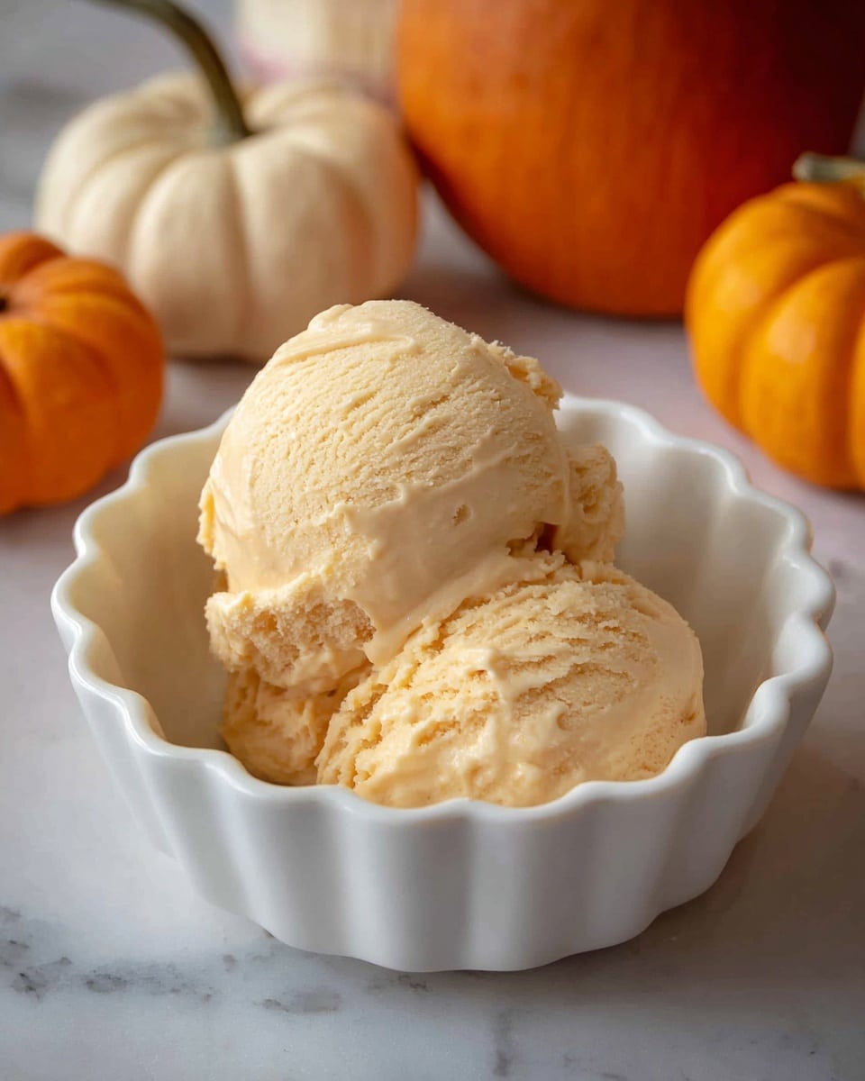 A fluted white bowl holds two scoops of creamy pumpkin ice cream, showing smooth, slightly textured surfaces with soft ridges and a pale orange color. The ice cream looks rich and dense with visible small air pockets. In the blurred background, there are small orange pumpkins and one white pumpkin placed on a white marbled surface. Photo taken with an iphone --ar 4:5 --v 7