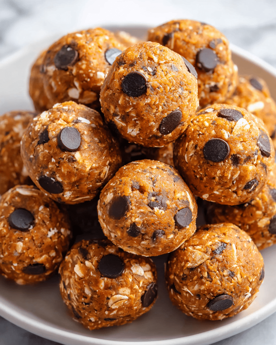 The image shows a close-up of a pile of small, round energy bites on a white plate. Each bite has a dense, coarse texture with visible oats embedded throughout their light brown base. Dark chocolate chips are scattered on the surface, contrasting with the lighter, slightly orange-brown color of the bites. The bites are tightly packed, showing their rounded shape and rough, uneven surface made up of oats and chocolate chips. The background is a white marbled texture. photo taken with an iphone --ar 4:5 --v 7