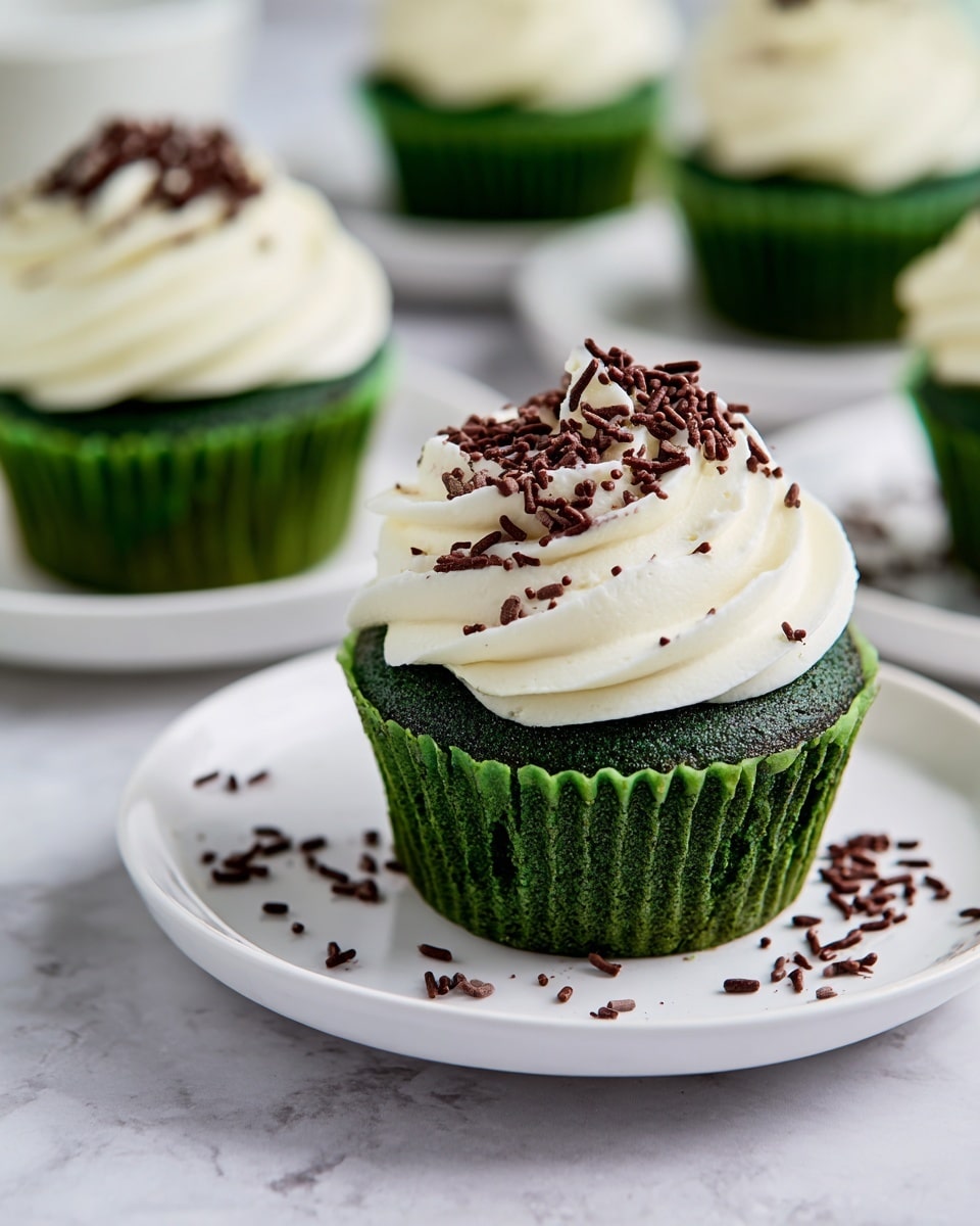 The image shows a close-up of green cupcakes with a swirl of white frosting on top. Each cupcake has a dark green base with a moist, slightly shiny texture, and thick white cream swirled in a smooth, round shape on top. The frosting is sprinkled with small dark brown chocolate sprinkles, some scattered on the white plate underneath. The cupcakes rest on simple white plates, all placed on a white marbled surface. The background is softly blurred with more cupcakes on plates visible. photo taken with an iphone --ar 4:5 --v 7