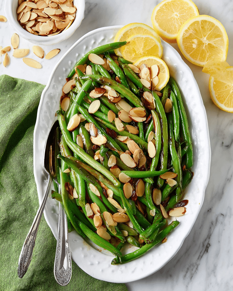 A white oval plate holds a fresh green bean dish on a white marbled surface. The dish has two layers: the bottom layer is long, cooked green beans with a shiny, smooth texture, stacked neatly but naturally. The top layer consists of toasted almond slices, light brown with some darker spots, scattered evenly over the green beans, adding a crunchy texture contrast. Around the plate are lemon halves and thin lemon slices, bright yellow with visible pulp, and a small white bowl on the side filled with more almond slices. A green cloth napkin partially enters from the bottom left, holding a silver fork with a detailed handle. Photo taken with an iphone --ar 4:5 --v 7