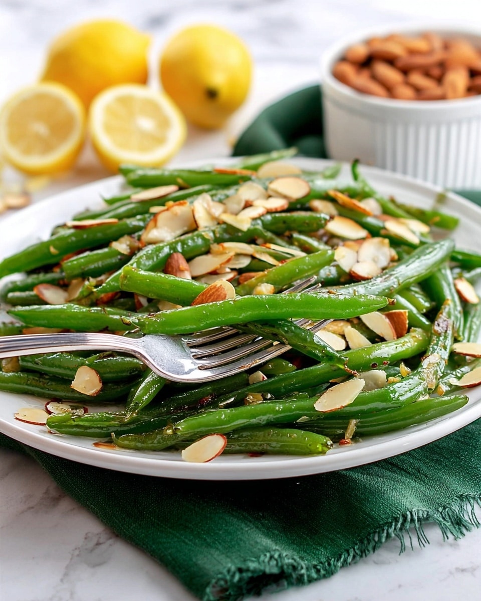 A white plate holds a generous serving of green beans topped with sliced, toasted almonds scattered evenly over the beans. The green beans shine slightly with a light coating of oil, showing some roasted spots in darker green to brown tones. A silver fork is holding a small bunch of the green beans in the foreground. In the background, there are lemon wedges, some almonds in a white bowl, and a green cloth napkin under the plate, all set on a white marbled surface. photo taken with an iphone --ar 4:5 --v 7