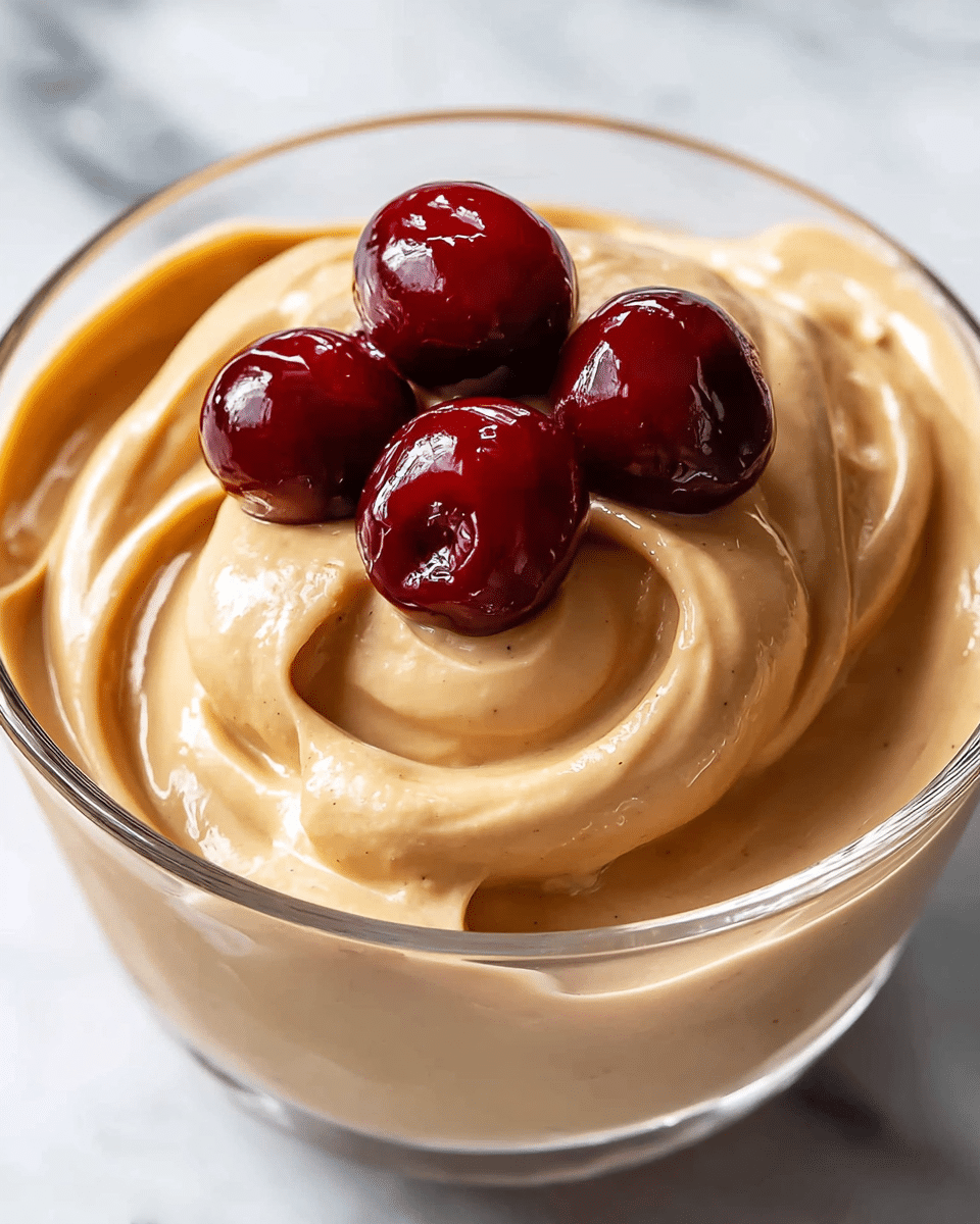 A close-up of a creamy light brown mousse in a clear glass cup, with a smooth, swirled texture forming soft peaks on the surface. On top, there are five shiny deep red cherries grouped in the center, adding a rich contrast to the light brown mousse below. The background is white marbled texture, highlighting the glossy and smooth look of the dessert. photo taken with an iphone --ar 4:5 --v 7