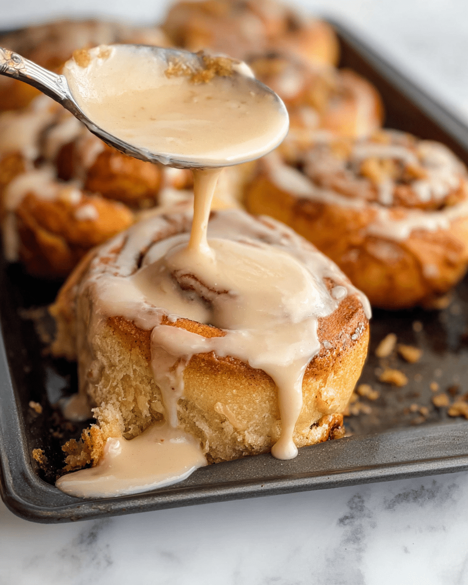 The image shows a close-up of a silver spoon pouring light brown, creamy glaze over a golden brown cinnamon roll in a dark baking tray. The cinnamon roll has a rough, textured surface with crispy edges and a soft, fluffy inside. The glaze is dripping down the sides of the roll, pooling slightly on the tray below, and covering other cinnamon rolls in the background with a smooth, shiny layer. The scene is set on a white marbled surface. photo taken with an iphone --ar 4:5 --v 7