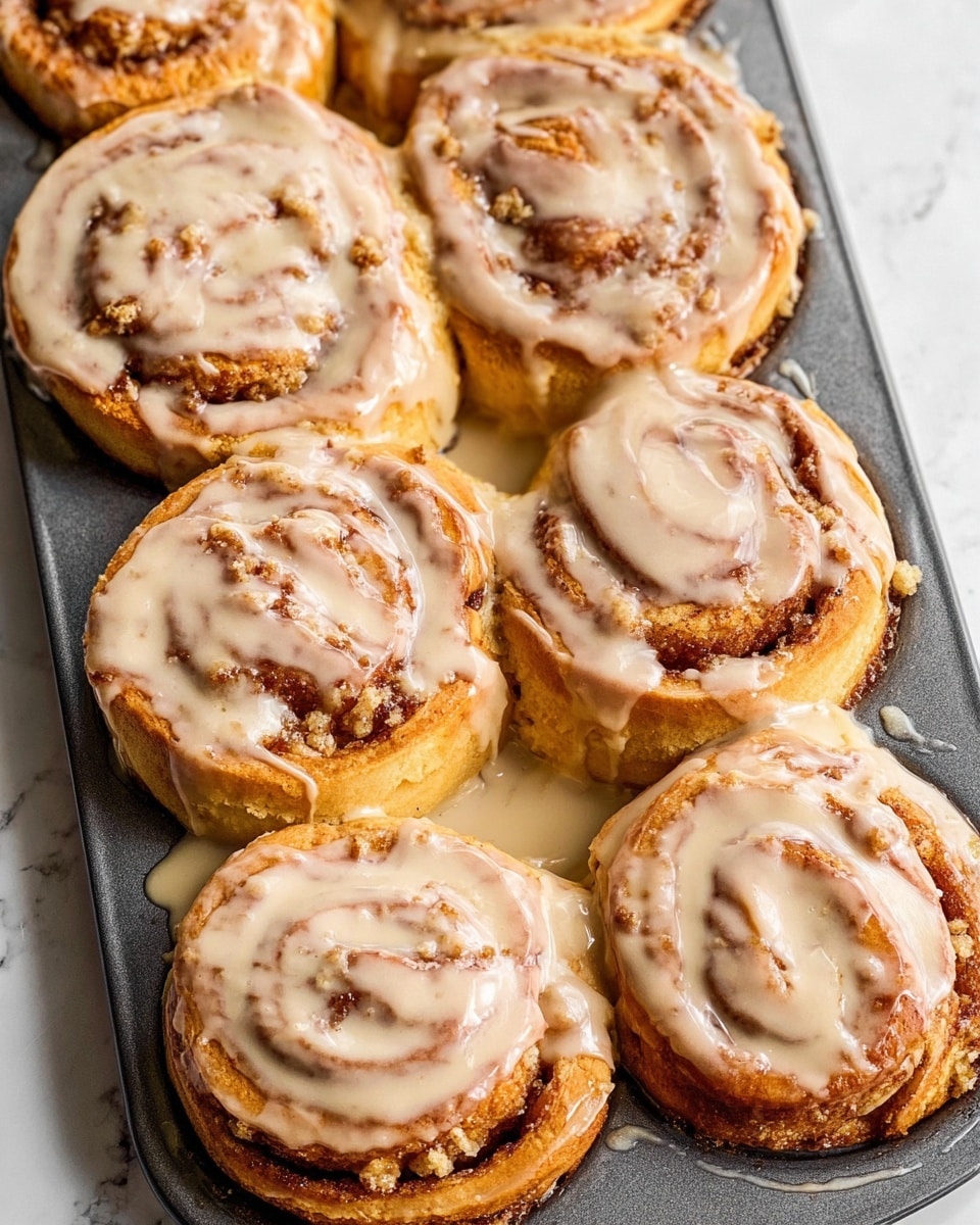 The image shows eight golden-brown cinnamon rolls placed closely together in a dark gray baking tray, each topped with a thick layer of creamy, light beige glaze that is smooth and shiny, dripping down their sides and pooling slightly in the tray. The cinnamon rolls have a rough, crumbly texture visible on top, suggesting a streusel or crumb topping beneath the glaze. The rolls are tightly coiled, showing layers of soft dough with darker cinnamon swirls inside. The tray is set against a white marbled background that enhances the warm tones of the cinnamon rolls. photo taken with an iphone --ar 4:5 --v 7