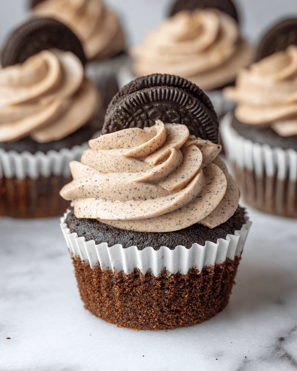 A close-up image shows a chocolate cupcake with three layers: a moist dark brown cake base in a white paper liner, topped with a thick swirl of light brown, speckled creamy frosting, all sitting on a white marbled surface. In the background, several more cupcakes are visible, each with a similar light brown frosting swirl and a dark chocolate cookie half placed vertically on top. The texture of the frosting looks smooth and fluffy, while the cake base appears soft and slightly crumbly. Photo taken with an iphone --ar 4:5 --v 7