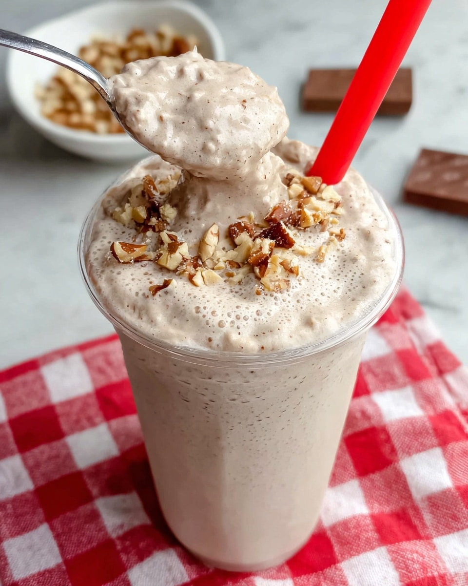 A tall clear plastic cup is filled with a creamy, light brown milkshake topped with small chunks of chopped nuts embedded in the thick foam layer. A shiny silver spoon is held above the cup, scooping up a dense, frothy portion of the milkshake with visible small nut pieces mixed in. A bright red straw peeks out from the cup. The cup sits on a red and white checkered cloth against a white marbled surface. In the background, a white bowl with more nut pieces is slightly out of focus along with a small piece of chocolate. Photo taken with an iphone --ar 4:5 --v 7