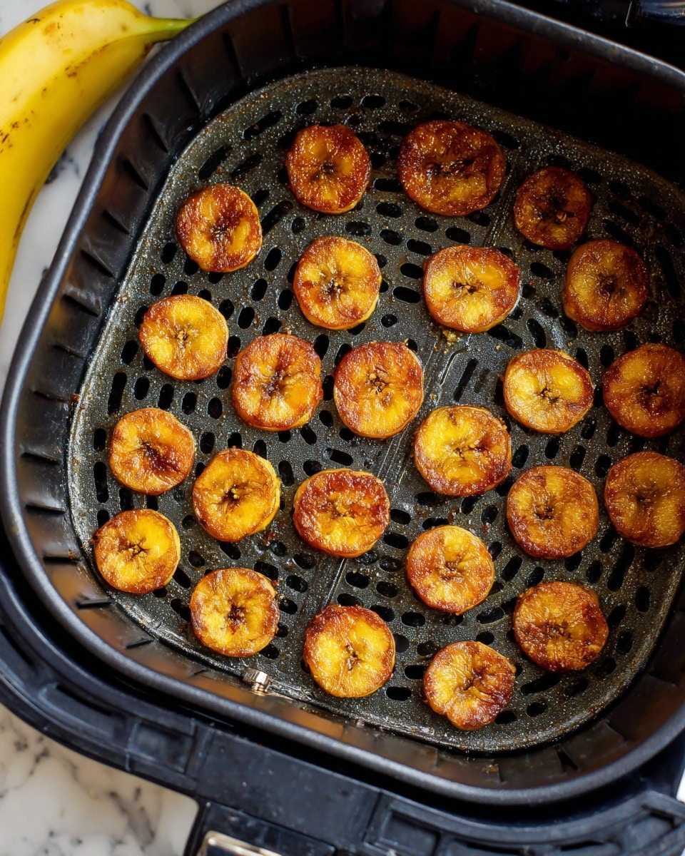Inside a black air fryer basket with round holes at the bottom, there are about twenty small golden-brown, round slices of cooked plantains arranged mostly in rows. Each plantain piece has a slightly crispy, caramelized surface with a mix of darker and lighter brown spots. The basket shows some light oil and seasoning residue. Part of a yellow banana and a white marbled surface are visible around the air fryer. photo taken with an iphone --ar 4:5 --v 7
