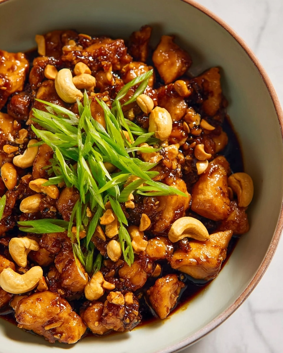 The image shows a close-up of a bowl filled with a rich, dark brown sauce coating bite-sized pieces of golden-brown cooked chicken. Mixed in are whole and halved light tan cashew nuts scattered throughout the dish. On top, there is a fresh garnish of bright green, thinly sliced scallions adding a fresh pop of color. The bowl is white, and the background features a white marbled texture. Photo taken with an iphone --ar 4:5 --v 7