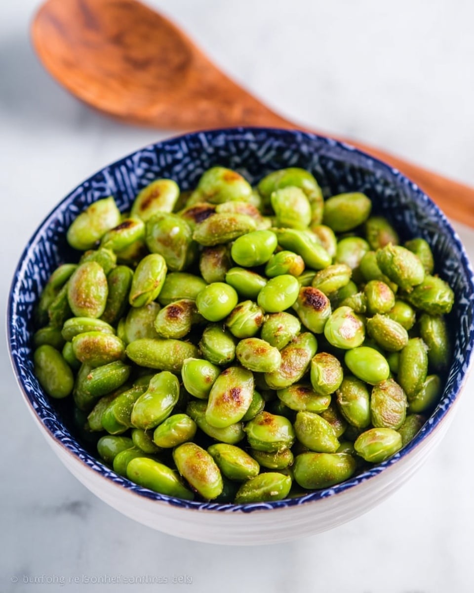 A close-up view of a bowl filled with roasted green edamame beans, some beans have a light brown charred texture on the surface, giving a crunchy look. The bowl is white with dark blue patterns around the rim and body. The bowl is placed on a white marbled surface. A wooden spoon with a smooth texture is partially visible behind the bowl. Photo taken with an iphone --ar 4:5 --v 7