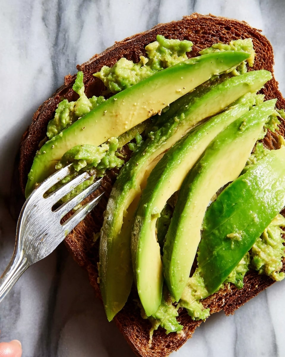 A close-up of one slice of dark brown toasted bread laid flat on a white marbled surface, topped with green avocado slices and mashed avocado spread on the left side. The avocado is cut into five uneven pieces, with the thicker slices on the right and mashed parts on the left, showing smooth and creamy textures mixed with some fork marks. A silver fork held by a woman's hand presses into the avocado, adding texture to the mash. photo taken with an iphone --ar 4:5 --v 7