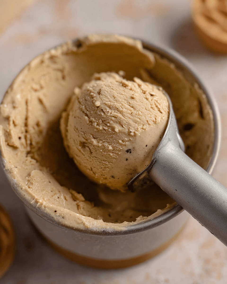 A close-up image shows a scoop of light brown ice cream with small dark specks held by a metallic ice cream scoop inside a round container. The ice cream appears creamy with a smooth texture and some slight roughness on the surface, and the container is nearly full with ice cream visible all around the scoop. The background features a white marbled texture with soft, out-of-focus objects, adding a warm, cozy feel to the scene. photo taken with an iphone --ar 4:5 --v 7