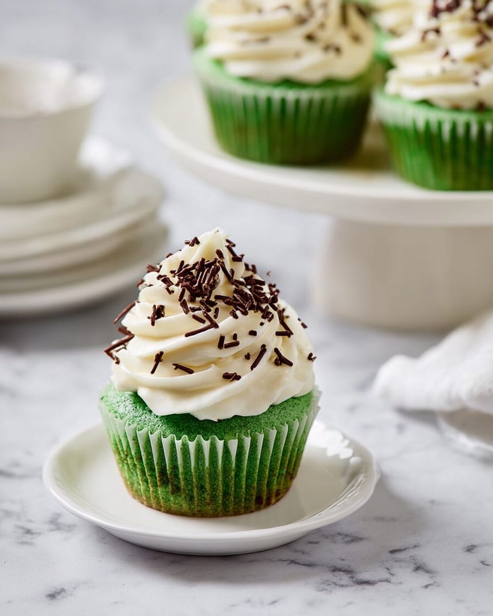 A green cupcake sits on a small white plate, topped with a thick swirl of white frosting that has a soft, creamy texture. The frosting is sprinkled lightly with thin, dark brown chocolate sprinkles that add contrast to the bright green and white colors. The cupcake’s green base looks moist and fluffy, taking up the bottom half of the cupcake, while the frosting forms a tall peak above it. Nearby, more green cupcakes with white frosting and similar chocolate sprinkles are placed on white plates and a white cake stand, all set on a white marbled surface. Photo taken with an iphone --ar 4:5 --v 7