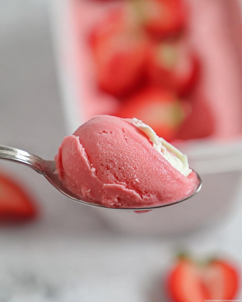 A close-up image of a metal spoon holding a smooth scoop of pink frozen dessert with a glossy, slightly creamy texture; a small part of white cream is visible behind the pink scoop on the spoon. The blurred background shows a white container with more of the pink dessert and a few pieces of sliced strawberries with red color and soft texture. The whole scene is set against a white marbled surface. Photo taken with an iphone --ar 4:5 --v 7