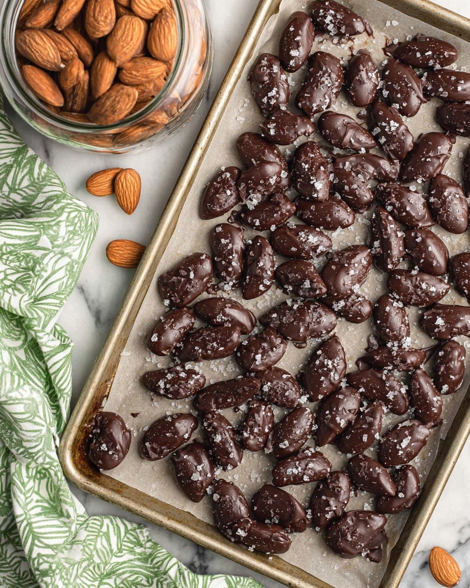 The image shows a metal tray lined with light parchment paper, covered with many dark chocolate-covered almonds sprinkled with coarse sea salt, each almond having a glossy, uneven texture with some chocolate crinkles. On the top left corner, a glass jar filled with whole almonds with smooth brown skin is partially visible. The bottom left corner has a green and white patterned cloth casually placed, with the whole scene set on a white marbled surface. Photo taken with an iphone --ar 4:5 --v 7