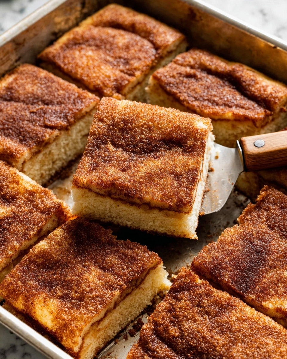 A close-up of nine square pieces of cinnamon coffee cake with a thick, golden brown layer of cinnamon sugar crust on top, each piece showing a soft, light beige cake layer underneath. One piece is being lifted by a spatula with a wooden handle, revealing the fluffy inside texture and the crispy top. The cake squares are arranged closely together in a metal baking tray with a shiny, slightly worn surface, all set on a white marbled texture. photo taken with an iphone --ar 4:5 --v 7