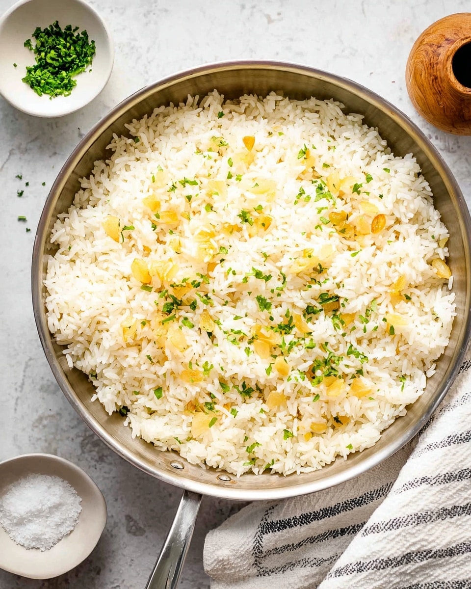 A close-up view of a skillet filled with a single layer of cooked white rice mixed with small yellow slices of fried garlic and sprinkled with chopped green herbs on top, sitting on a white marbled textured surface. To the upper left of the skillet is a small white bowl with finely chopped herbs, and a wooden container with coarse salt is nearby. A white cloth with thin black stripes is placed to the right side of the skillet, partially under the handle. The rice looks soft with a light fluffy texture, and the garlic slices add a crisp, golden contrast. Photo taken with an iphone --ar 4:5 --v 7