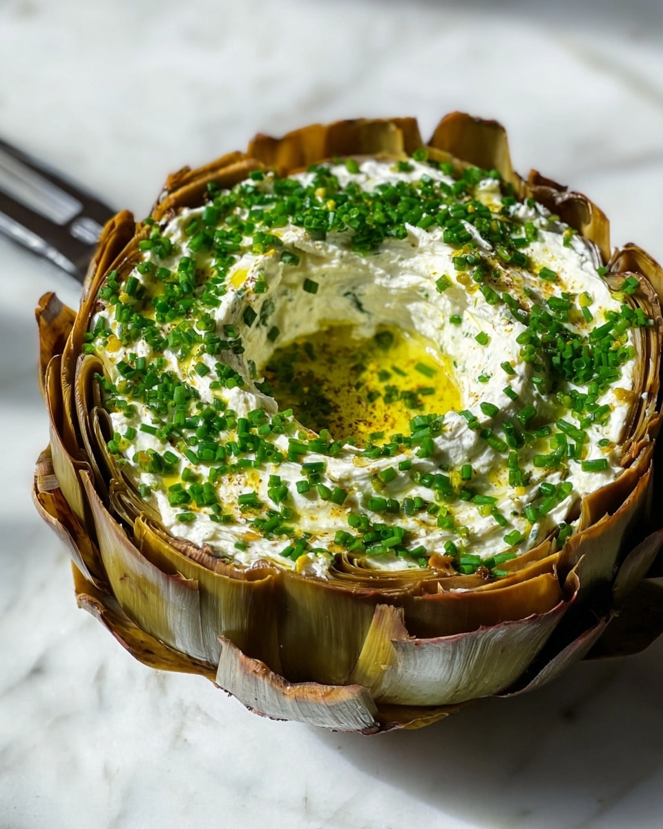 The dish shows a layered presentation starting with an outer ring of brownish artichoke leaves, tightly wrapped and forming the base layer. Inside this is a thick, creamy white spread mixed with green herbs, making the middle layer soft and smooth. The top layer is generously covered with finely chopped bright green chives scattered around, adding color contrast and texture. The creamy layer has a small hollow in the center, filled with a drizzle of golden olive oil that shines under the light. The dish is placed on a white marbled surface, giving a clean and elegant look, with a silver kitchen tool visible on the side. Photo taken with an iphone --ar 4:5 --v 7