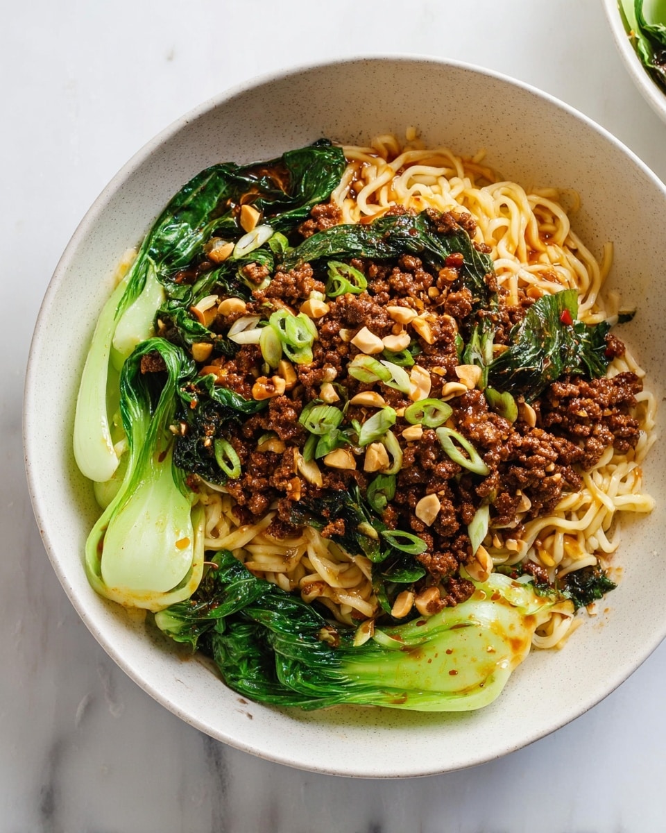 A white bowl holds a dish with three main layers. The bottom layer is bright green bok choy leaves with light green stems, arranged around the edges. Above that is a thick pile of pale yellow noodles, round and slightly glossy. The top layer is a mix of browned ground meat and dark green cooked leafy vegetables, scattered unevenly. Small chopped peanuts and light green sliced scallions are sprinkled on top, adding texture and color contrast. A light brown sauce is drizzled lightly over the dish. The bowl is placed on a white marbled surface. Photo taken with an iphone --ar 4:5 --v 7